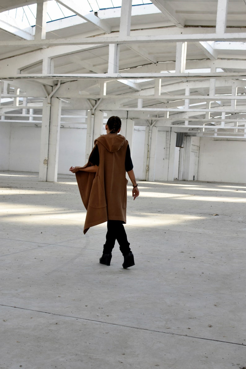 May include: A person wearing a brown hooded vest with a long, flowing design. The vest has a relaxed fit and is worn over a black shirt. The person is standing in a large, empty room with a white ceiling.