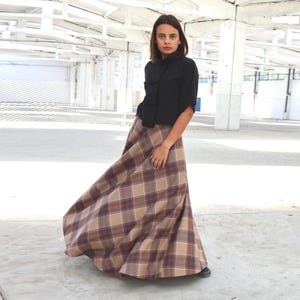 Pode incluir: Uma blusa preta com gola alta e uma saia longa xadrez fluida em tons de castanho, bege e bordô. A saia tem um design amplo e rodado. A roupa é usada em um espaço claro e aberto.