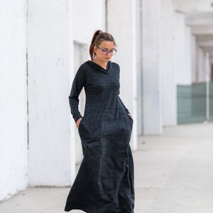 May include: A woman wearing a long, dark gray hooded dress with large pockets. She is standing in a white hallway and wearing black sneakers.