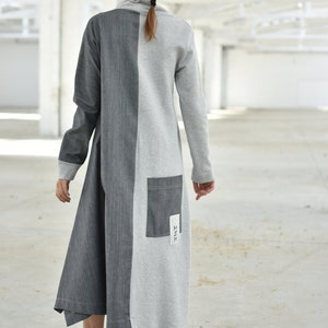 Puede incluir: Un vestido largo, gris y gris oscuro con un panel contrastante en el centro. El vestido tiene cuello alto y mangas largas. El vestido tiene un peque&ntilde;o bolsillo en la espalda con la palabra "HUSH" escrita en &eacute;l.