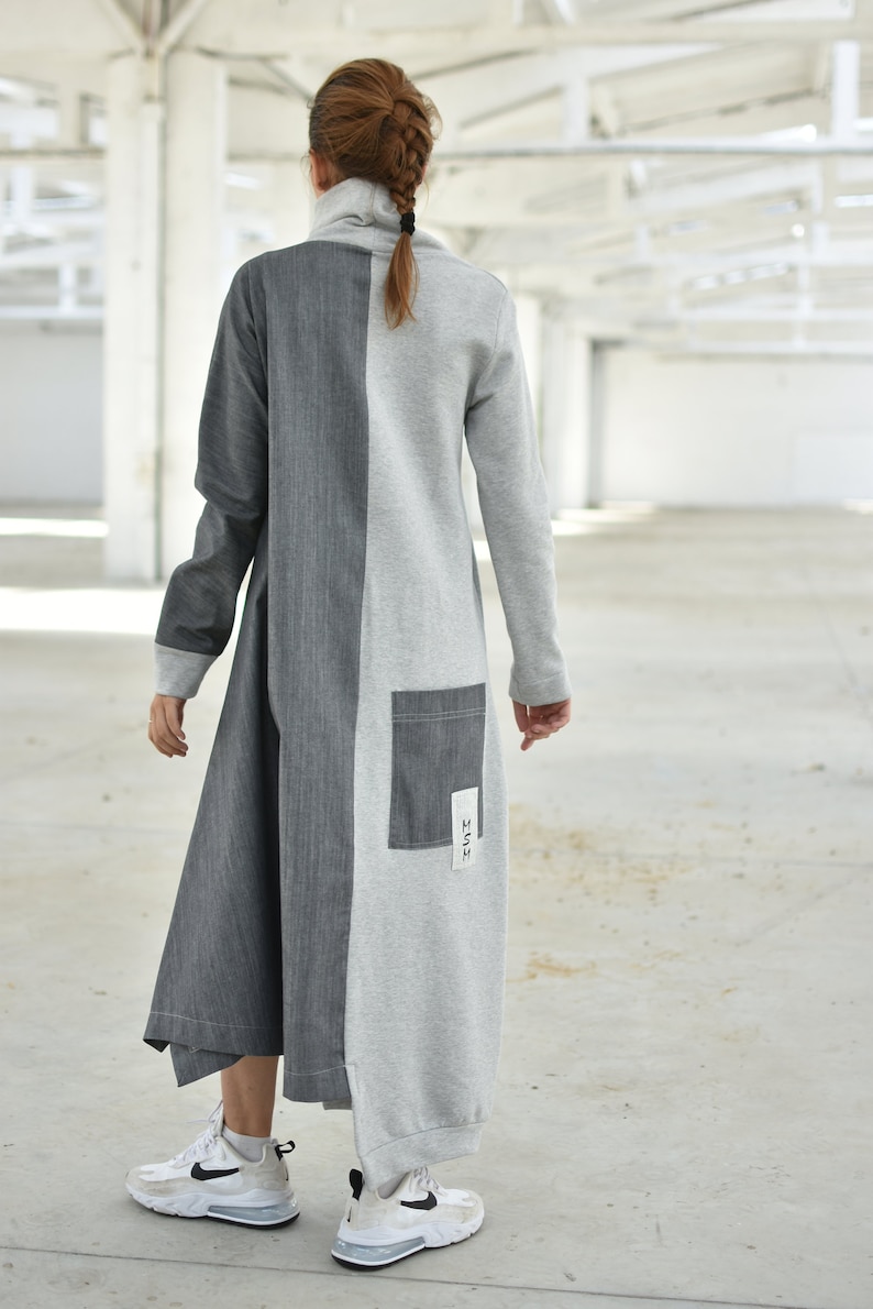Puede incluir: Un vestido largo, gris y gris oscuro con un panel contrastante en el centro. El vestido tiene cuello alto y mangas largas. El vestido tiene un peque&ntilde;o bolsillo en la espalda con la palabra "HUSH" escrita en &eacute;l.