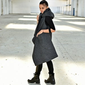May include: A long, gray hooded vest with a draped design. The vest has a large hood and pockets. The model is wearing black pants and black boots.