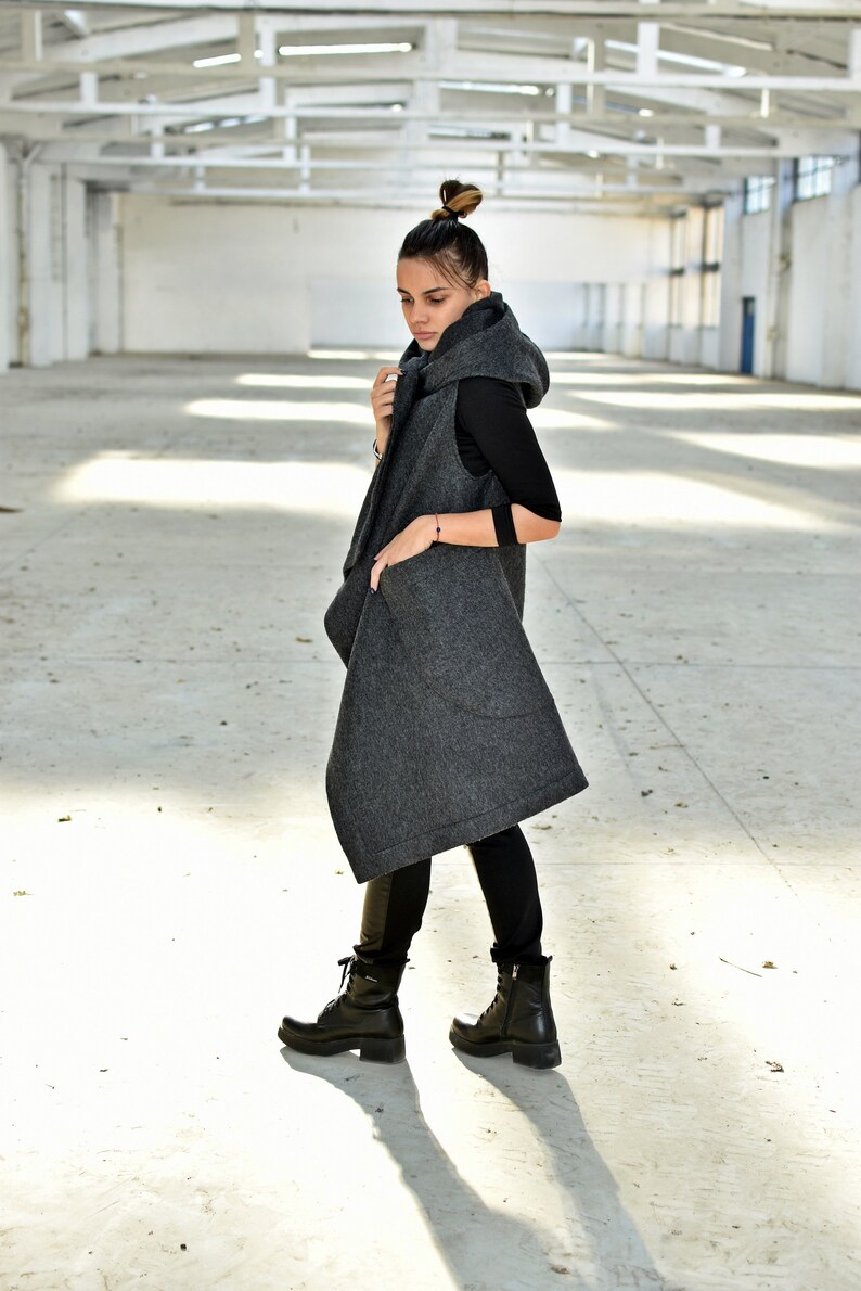 May include: A long, gray hooded vest with a draped design. The vest has a large hood and pockets. The model is wearing black pants and black boots.