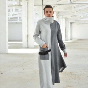 Puede incluir: Un vestido largo de manga larga gris y negro con cuello alto y cintur&oacute;n negro. El vestido tiene un dise&ntilde;o &uacute;nico con un panel gris en el lado izquierdo y un panel negro en el lado derecho. La modelo lleva zapatillas blancas.