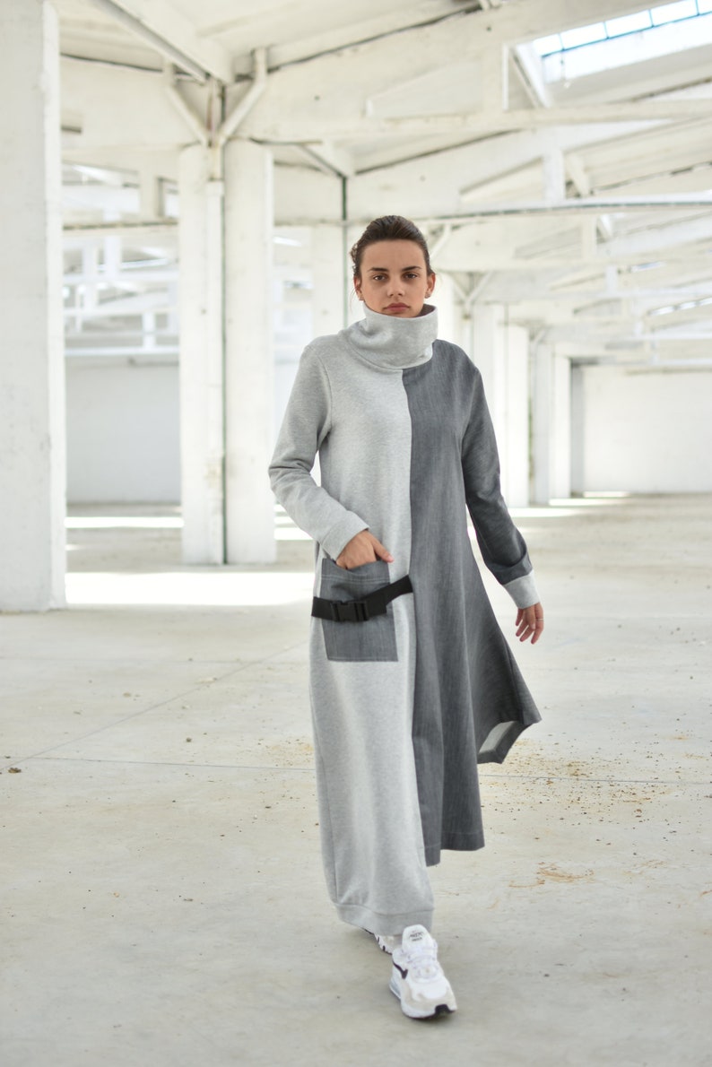 Puede incluir: Un vestido largo de manga larga gris y negro con cuello alto y cintur&oacute;n negro. El vestido tiene un dise&ntilde;o &uacute;nico con un panel gris en el lado izquierdo y un panel negro en el lado derecho. La modelo lleva zapatillas blancas.