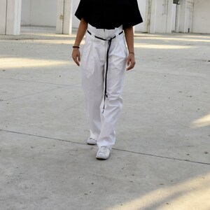 May include: A person wearing white pants with a black belt and white sneakers. The pants have pleats and a high waist.