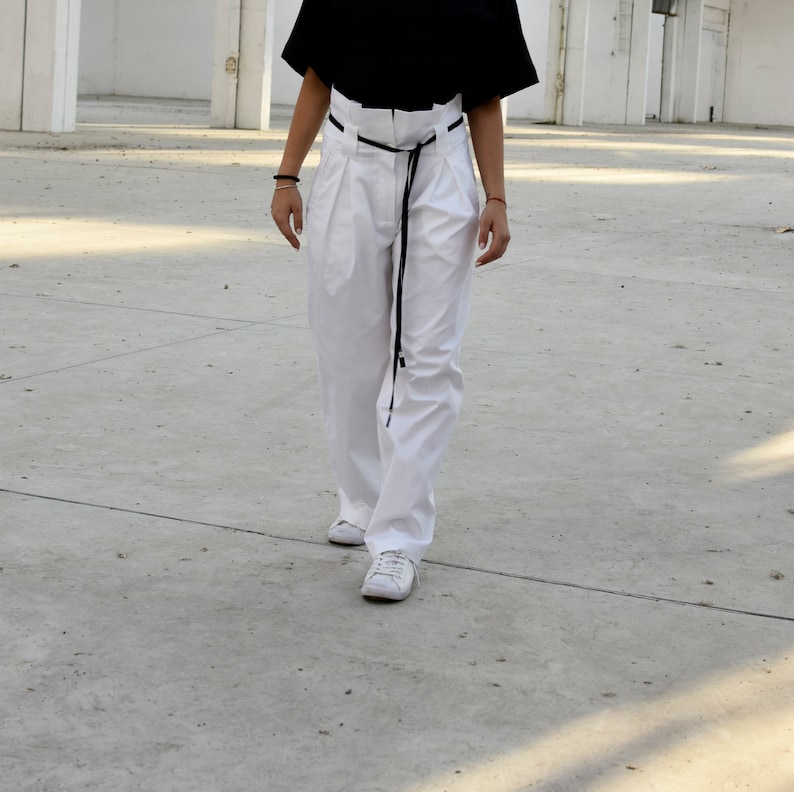 May include: A person wearing white pants with a black belt and white sneakers. The pants have pleats and a high waist.