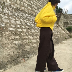 May include: A person wearing a bright yellow puffer jacket and wide-leg brown pants. The person is wearing black sneakers with blue accents. The background features a stone wall and a concrete walkway.