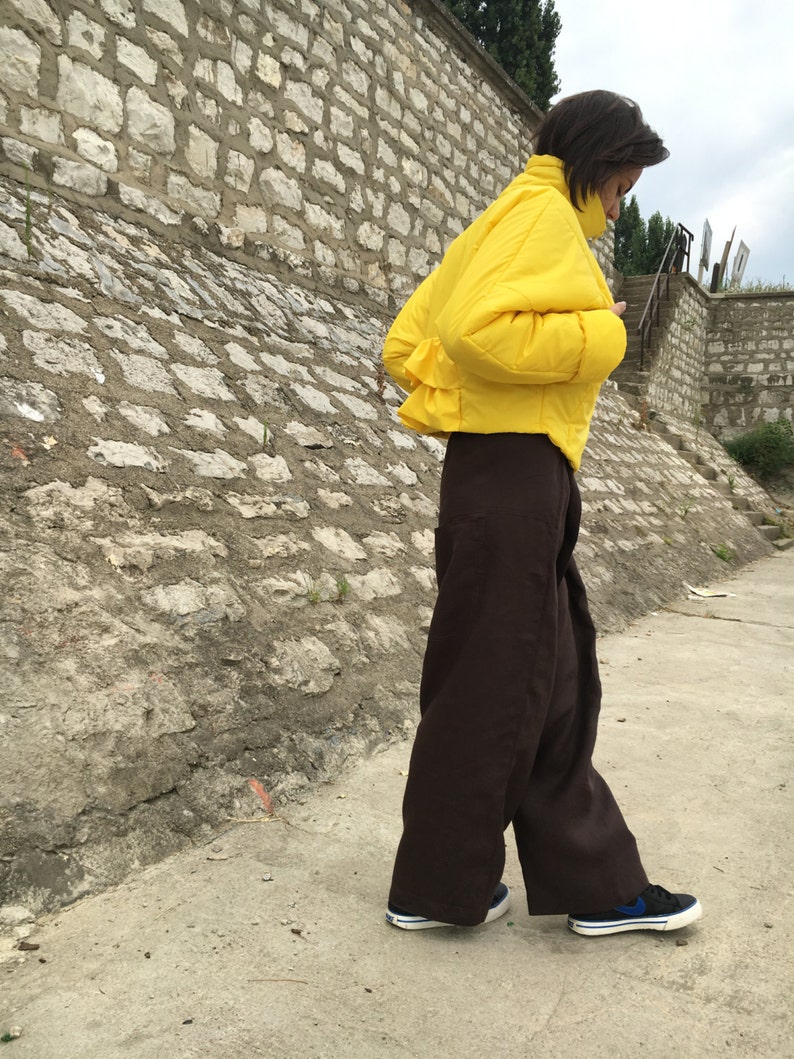 May include: A person wearing a bright yellow puffer jacket and wide-leg brown pants. The person is wearing black sneakers with blue accents. The background features a stone wall and a concrete walkway.