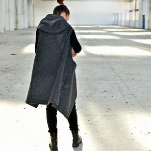 May include: A person wearing a gray hooded vest with a black shirt and black pants. The person is standing in a large, empty warehouse with white walls and a metal roof.