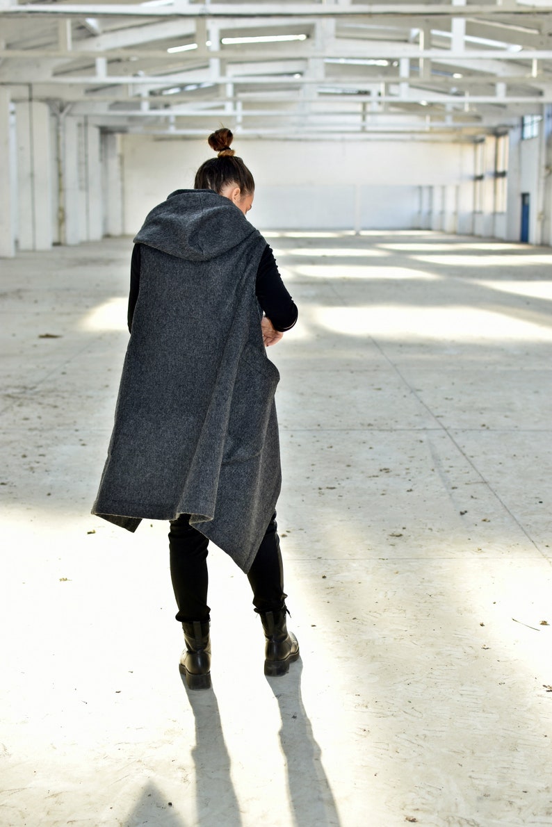May include: A person wearing a gray hooded vest with a black shirt and black pants. The person is standing in a large, empty warehouse with white walls and a metal roof.