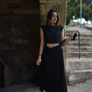 May include: A woman wearing a black crop top and a long black skirt with a side tie. She is standing in front of a stone wall and holding a black strap in her hand. She is wearing white sneakers.