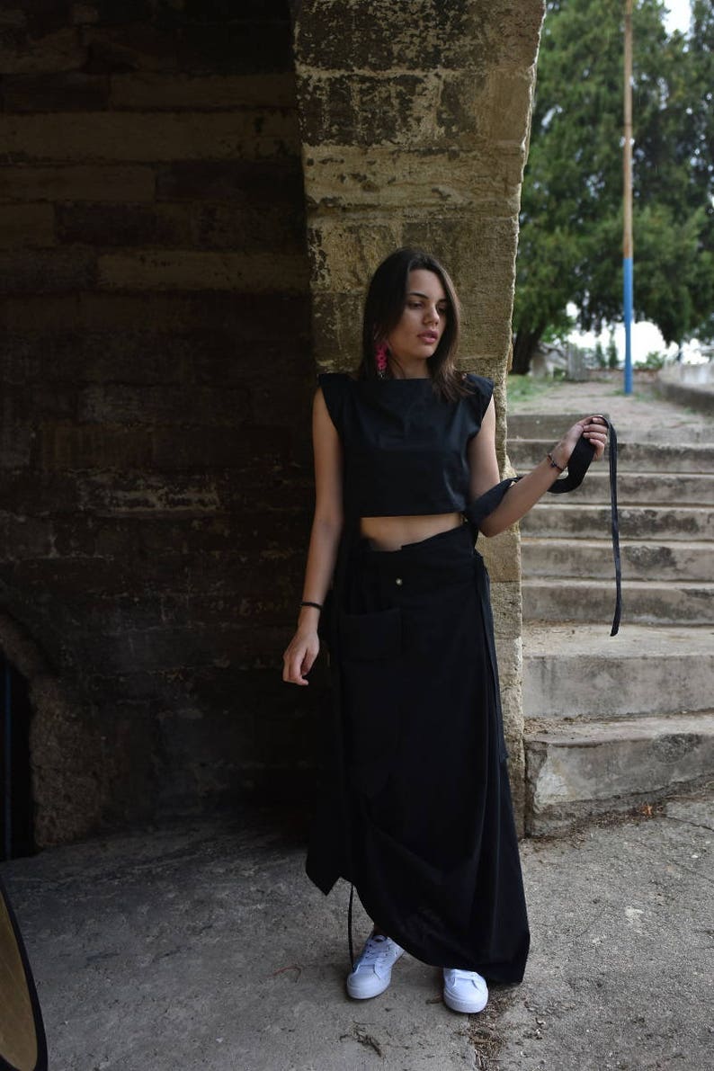 May include: A woman wearing a black crop top and a long black skirt with a side tie. She is standing in front of a stone wall and holding a black strap in her hand. She is wearing white sneakers.