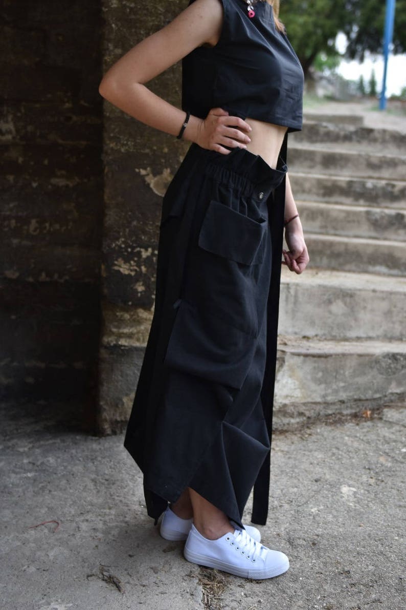 May include: A black sleeveless crop top and a black, long, flowing skirt with a large cargo pocket. The skirt has a drawstring waist and a belt. The person is wearing white sneakers.