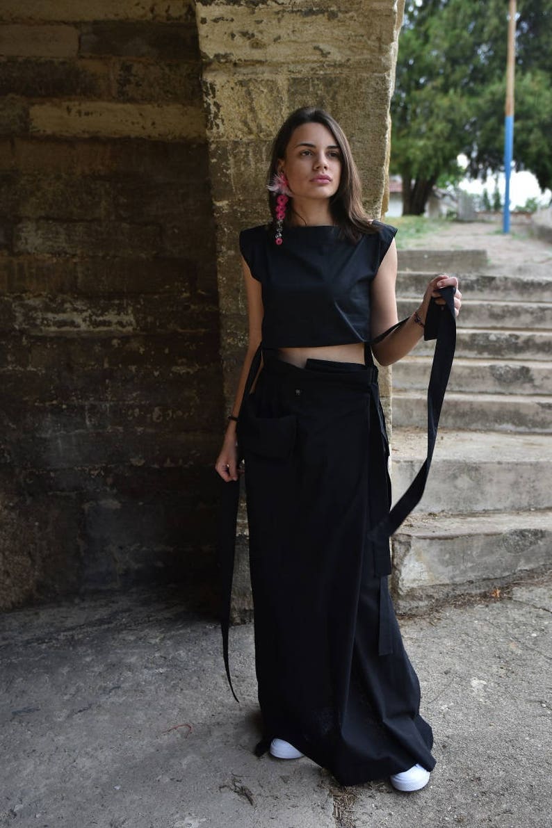 May include: A woman wearing a black crop top with a black maxi skirt and white sneakers. The top has a tie detail at the waist and the skirt has a large pocket on the side. She is wearing pink flower earrings.