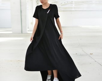 May include: A black short-sleeved maxi dress with a round neckline and an asymmetrical hemline. The dress features a draped front panel and a small white embroidered logo on the upper chest area. The garment is paired with white athletic sneakers.