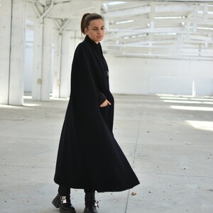 May include: A woman wearing a black cape coat with a high collar and a long, flowing silhouette. She is standing in a white, industrial setting.