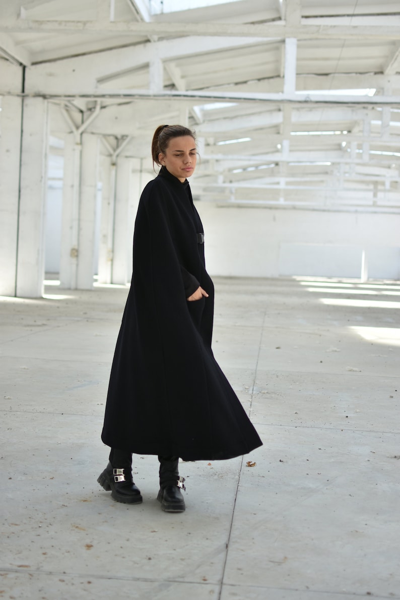 May include: A woman wearing a black cape coat with a high collar and a long, flowing silhouette. She is standing in a white, industrial setting.
