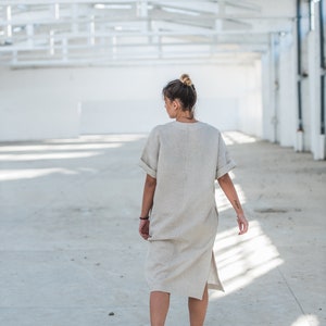 May include: A woman wearing a light beige linen dress walks in a white industrial space. The dress has a relaxed fit and a side slit. She is wearing white sneakers.