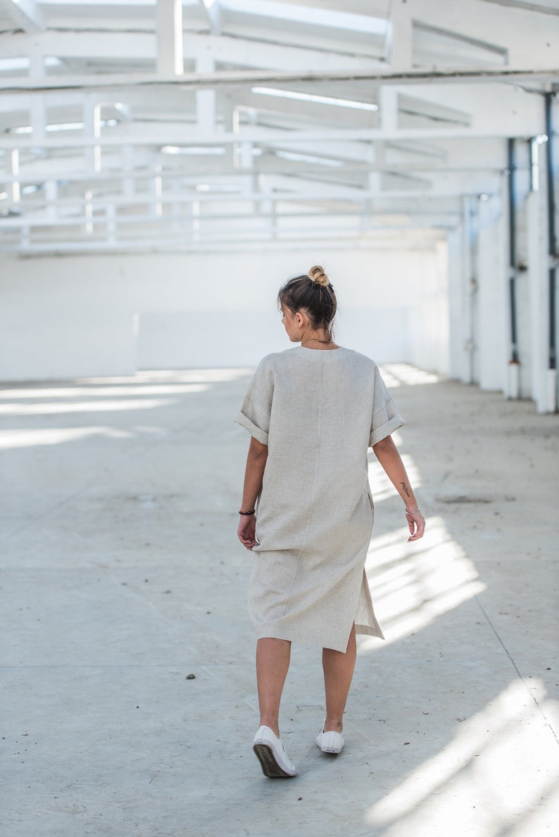 May include: A woman wearing a light beige linen dress walks in a white industrial space. The dress has a relaxed fit and a side slit. She is wearing white sneakers.