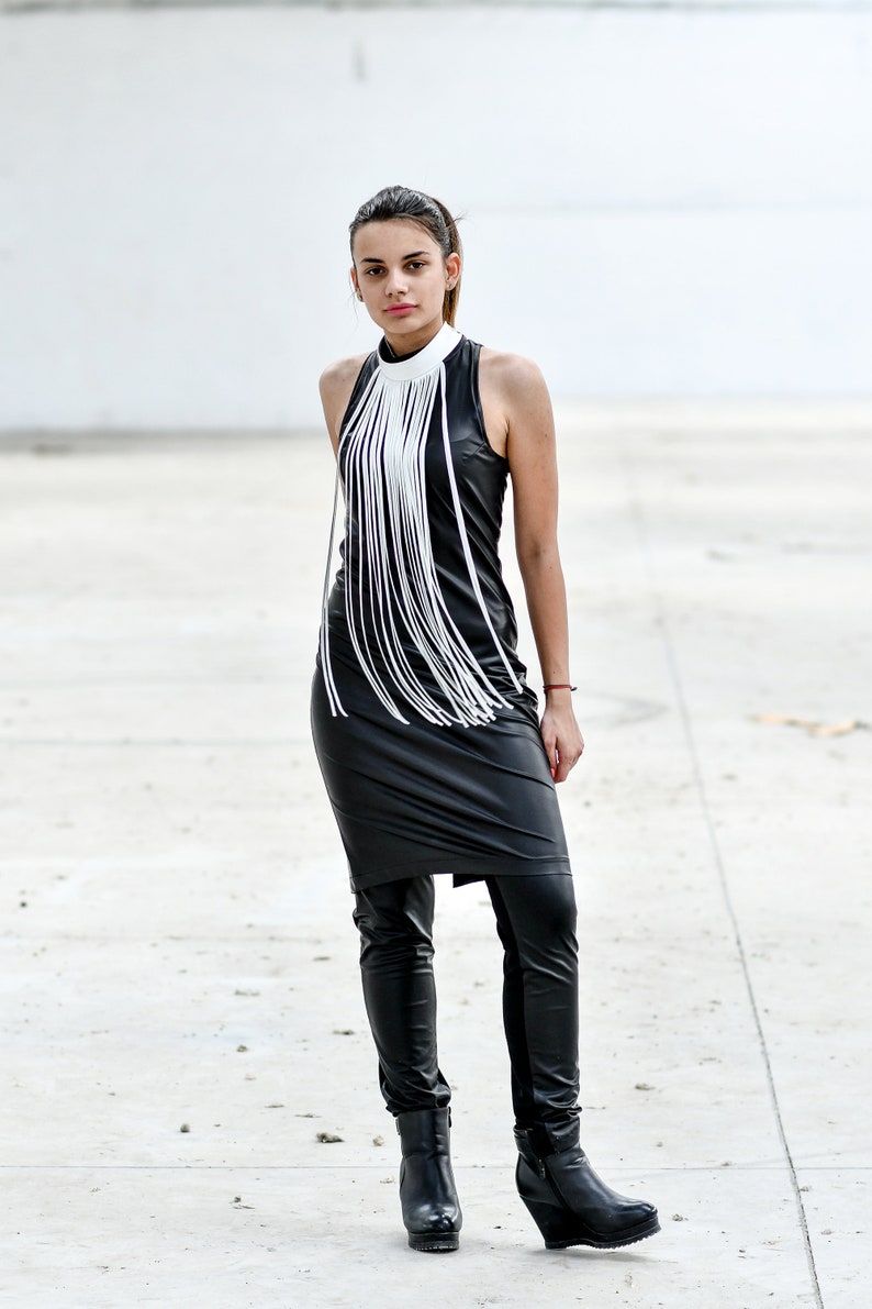 May include: A woman wearing a black leather dress with a white fringe detail at the neckline. She is also wearing black leather trousers and black boots.