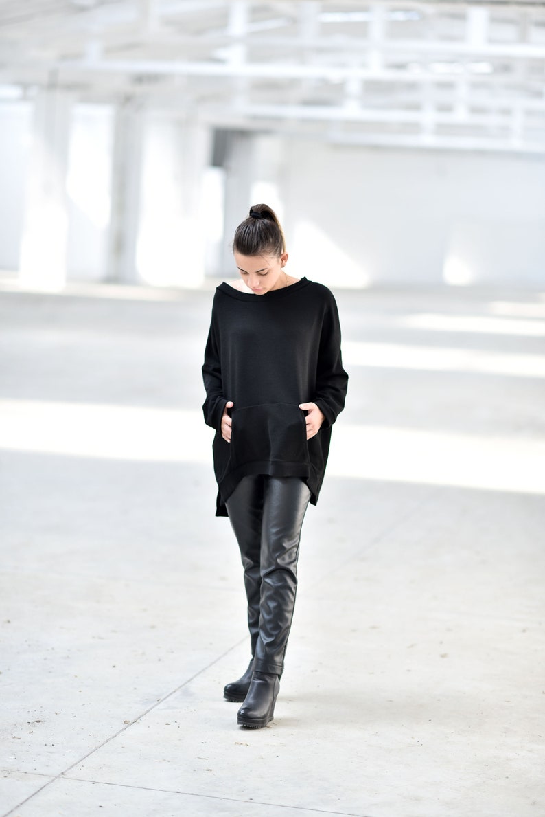 May include: A woman wearing a black oversized sweatshirt with a kangaroo pocket and black leather pants. She is standing on a concrete floor.