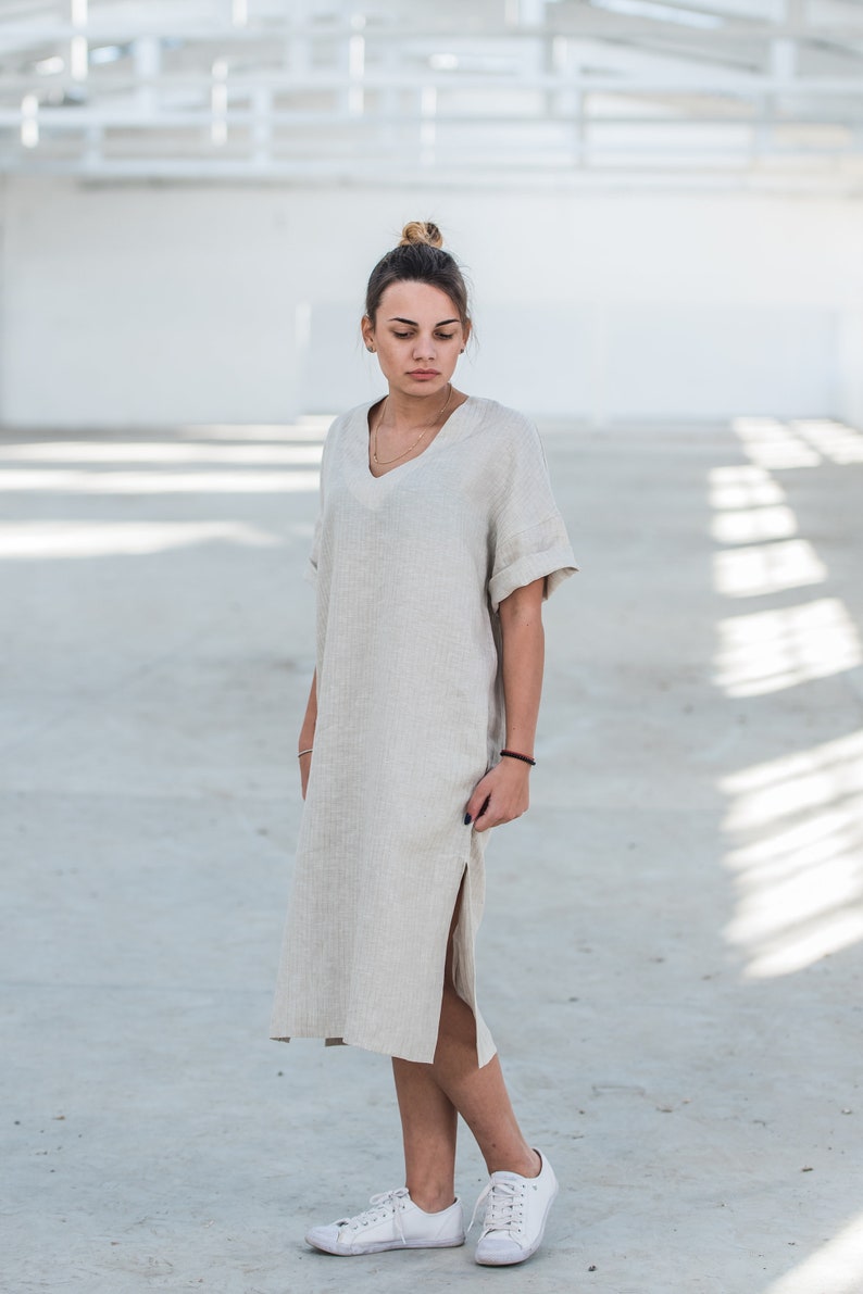 May include: A woman wearing a light beige linen dress with a side slit and short sleeves. She is wearing white sneakers and has her hair pulled back in a bun.