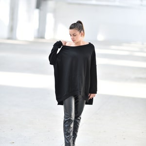 May include: A woman wearing a black oversized sweater with a pocket and black leather pants. She is standing in a white, industrial setting.