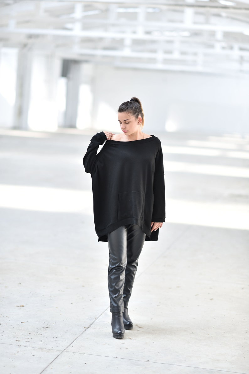 May include: A woman wearing a black oversized sweater with a pocket and black leather pants. She is standing in a white, industrial setting.