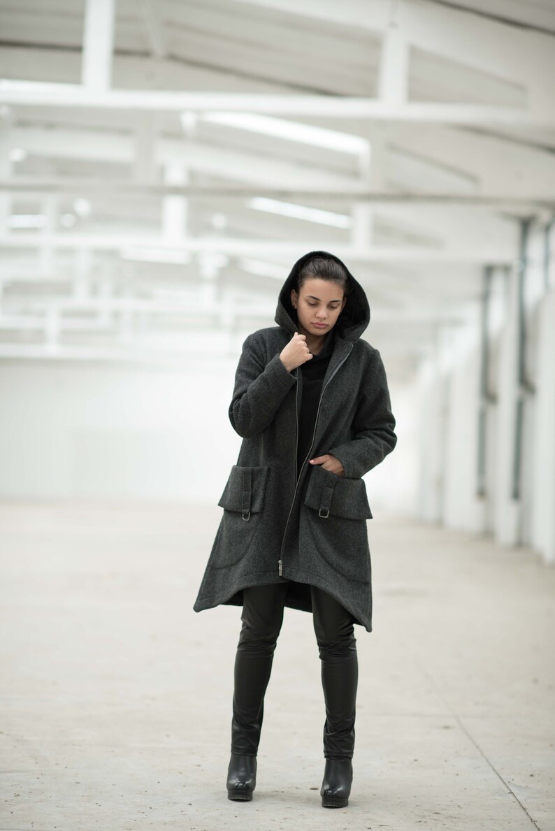 May include: A woman wearing a dark gray hooded coat with a zipper closure and two large pockets. She is wearing black pants and black boots.