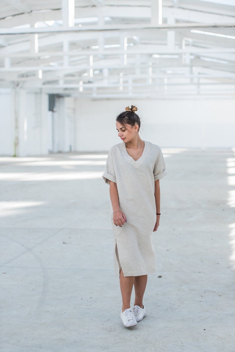 May include: A woman wearing a light beige linen dress with a v-neck and short sleeves. She is wearing white sneakers and has her hair pulled back in a bun.