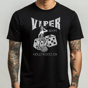 May include: Black t-shirt with a distressed white graphic. The graphic includes the text "VIPER ROOM" and an illustration of a mermaid-like figure on dice. The text "HOLLYWOOD CA" is below the dice.