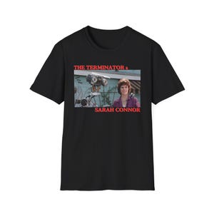 Może przedstawiać: Czarny T-shirt z grafiką przedstawiającą Sarah Connor i Terminatora z filmu Terminator 2: Dzień sądu. Tekst "THE TERMINATOR & SARAH CONNOR" jest nadrukowany na koszulce na czerwono.