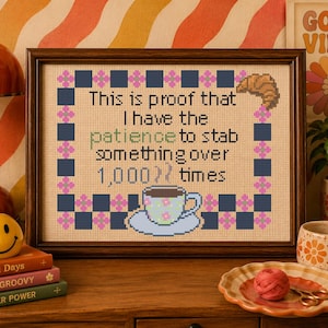 May include: A framed cross-stitch embroidery featuring the text "This is proof that I have the patience to stab something over 1,000 times" in black and green lettering, surrounded by a checkered border with a croissant and a coffee cup.