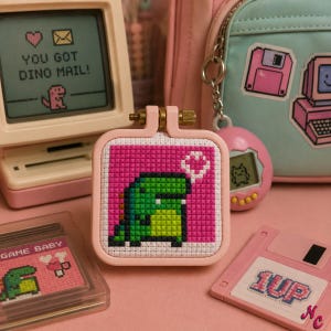 May include: A square embroidery hoop frame containing a cross-stitch pattern of a green pixelated dinosaur on a bright pink background. The dinosaur is facing right with a white heart shape above its head. The frame is light pink with a brass screw.