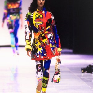May include: A model walks the runway wearing a colorful, patterned jacket and leggings. The jacket has a red, yellow, blue, and black pattern with a variety of graphic designs. The leggings are a similar pattern with a blue, yellow, and black color scheme. The model is carrying a small, colorful handbag.