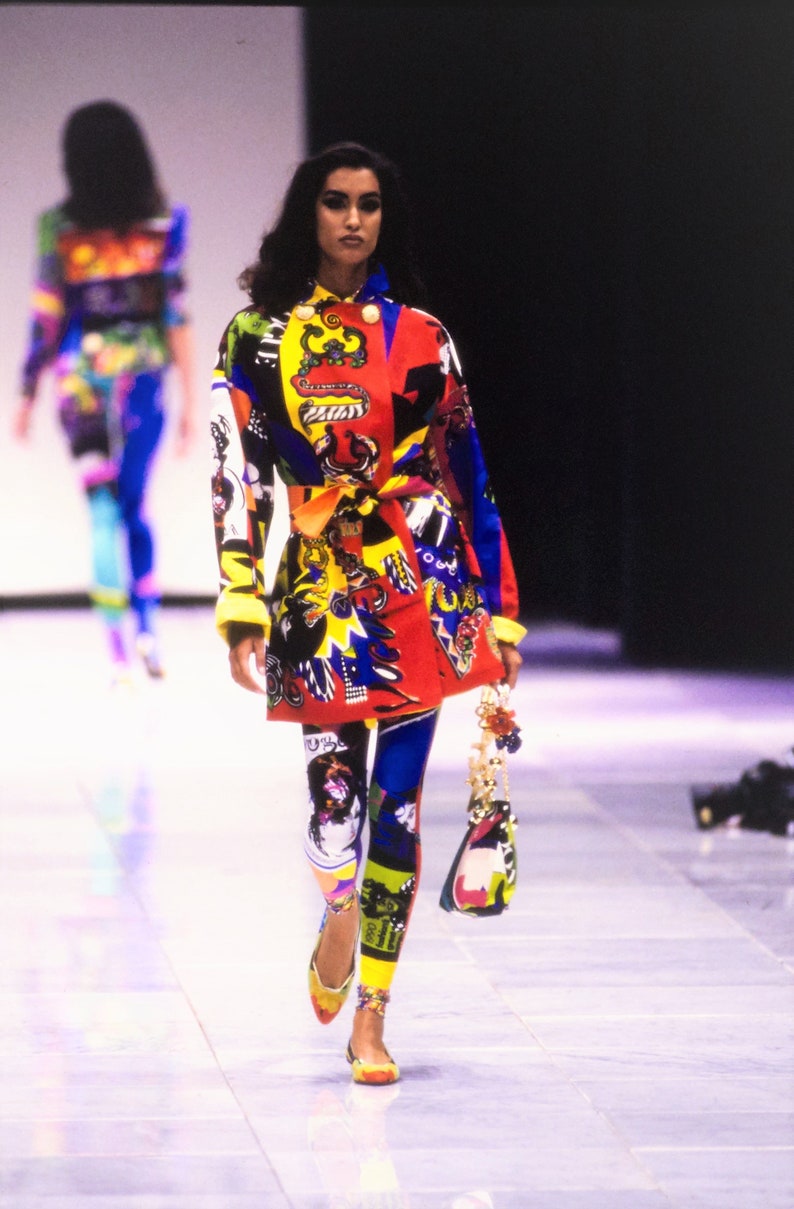May include: A model walks the runway wearing a colorful, patterned jacket and leggings. The jacket has a red, yellow, blue, and black pattern with a variety of graphic designs. The leggings are a similar pattern with a blue, yellow, and black color scheme. The model is carrying a small, colorful handbag.