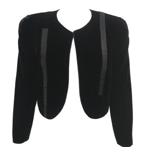 May include: A black velvet cropped jacket with long sleeves and a rounded neckline. The jacket has a satin trim along the edges.