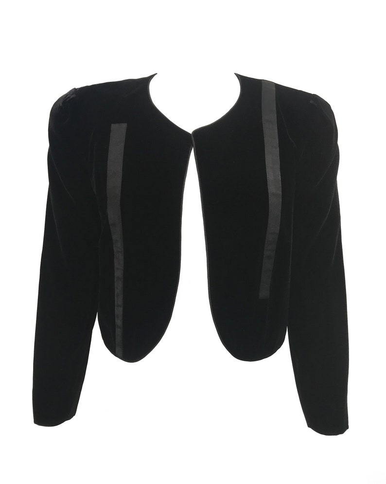 May include: A black velvet cropped jacket with long sleeves and a rounded neckline. The jacket has a satin trim along the edges.