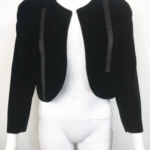 May include: A black velvet cropped jacket with satin trim. The jacket has a round neckline and long sleeves.
