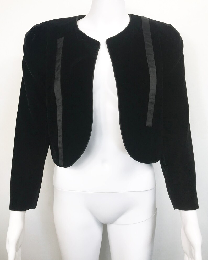May include: A black velvet cropped jacket with satin trim. The jacket has a round neckline and long sleeves.