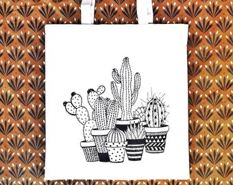 May include: White canvas tote bag with black line drawings of cacti in pots. The cacti are in various shapes and sizes, including a tall cactus with spines, a round cactus with spines, and a prickly pear cactus.