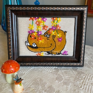 May include: A framed embroidery of a brown hippopotamus with a smiling face, surrounded by yellow, orange, pink, and purple flowers. The embroidery is on a white linen background.
