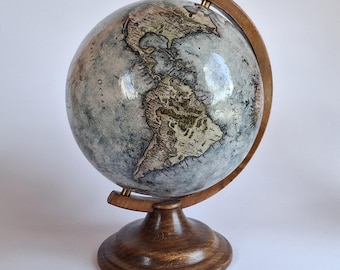 Op de afbeelding: Een globe in vintage-stijl met een gedetailleerde wereldkaart. De globe heeft een lichtblauw en grijs kleurenschema met bruine houten accenten. Hij is gemonteerd op een houten standaard en heeft een houten meridiaanring.