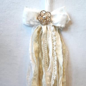 以下が含まれることがあります： クリーム色の生地のリボンに、金とパールの花の装飾。クリームとアイボリーの色合いの長いほつれた生地のストリーマー、繊細なレースのトリム付き。ヴィンテージ風の装飾品です。
