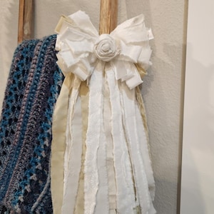 以下が含まれることがあります： 中央にバラのディテールと長いフリンジリボンが付いた、装飾用の白い布製リボン。リボンは木のダボに取り付けられており、背景には青と白のかぎ針編みの毛布が見えます。
