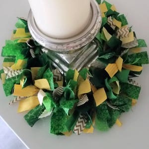 May include: A white candle sits on a silver candle holder surrounded by a green and yellow fabric wreath.