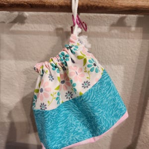 May include: A small drawstring bag with a floral pattern in pink, blue, and green on a white background. The bottom half of the bag is turquoise with a zebra-like pattern. The bag is suspended by a white cord and a pink metal clip.