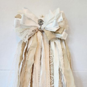 以下が含まれることがあります： クリーム色の、重ねられた布製のリボンで、長く流れるリボンが付いています。リボンにはレースと銀色のバラの装飾が施されています。リボンはクリーム色、ベージュ色、レースの組み合わせで、ヴィンテージでロマンチックな美しさを醸し出しています。