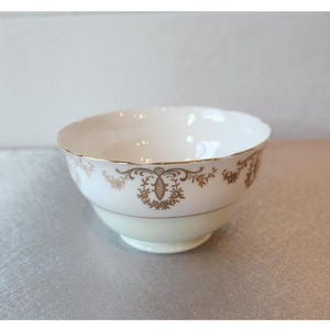 May include: A white porcelain bowl with a gold rim and decorative gold floral and geometric patterns. The bowl has a light cream-colored base and a scalloped edge. The design is symmetrical and elegant.